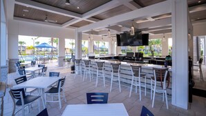 A covered outdoor seating area with tables and chairs, a bar, and a pool in the background.
