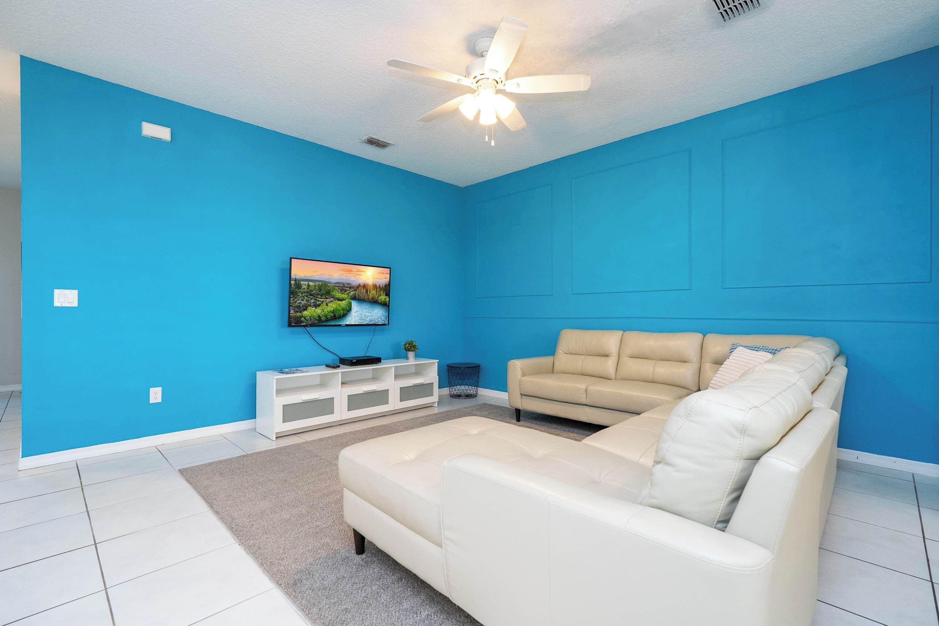 A living room with a blue wall, white ceiling fan, beige sofa, and a flat-screen TV mounted on the wall.