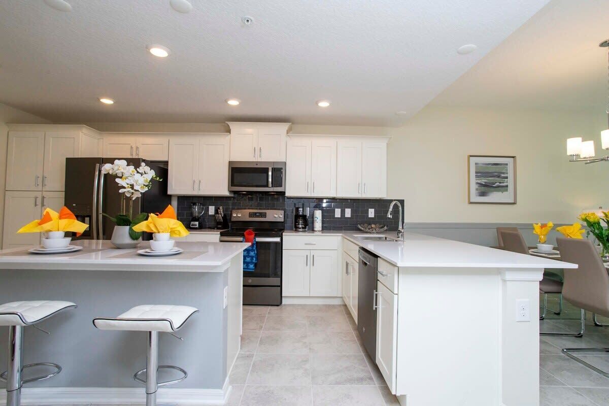 A modern kitchen with white cabinets, a central island with bar stools, a stainless steel refrigerator, a microwave, and an oven.