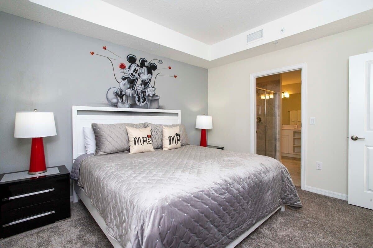 A bedroom with a large bed, two bedside tables with lamps, a white headboard, and a wall mural.