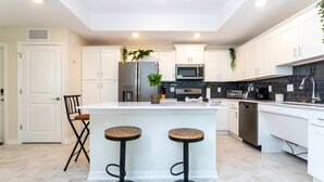 A modern kitchen with white cabinets, a central island with stools, stainless steel appliances, and a black tiled backsplash.
