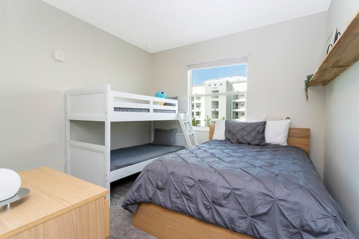 A bedroom with a bunk bed, a single bed, a desk, and a window with a view of buildings.