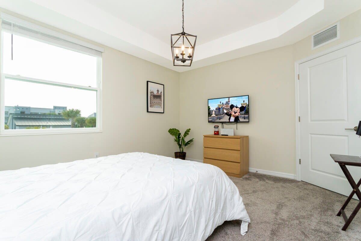 Een slaapkamer met een groot bed, een televisie op een standaard, een dressoir, een plant en een raam met uitzicht op gebouwen.