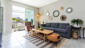 A modern living room with a grey sofa, a wooden coffee table, and a dining area with chairs. There are decorative mirrors and plants on the walls.