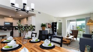 A modern kitchen with white cabinets, a dining area with a wooden table set for four, and a living area with a sofa and a TV.