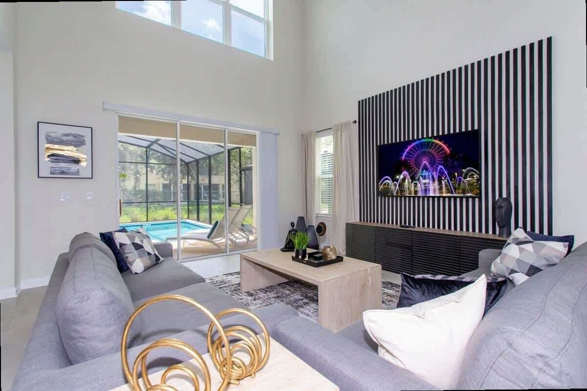 Uma sala de estar moderna com uma televisão grande, sofás cinza, uma mesa de centro e uma porta de vidro que dá para uma piscina.