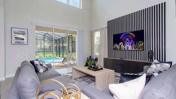 Uma sala de estar moderna com uma televisão grande, sofás cinza, uma mesa de centro e uma porta de vidro que dá para uma piscina.