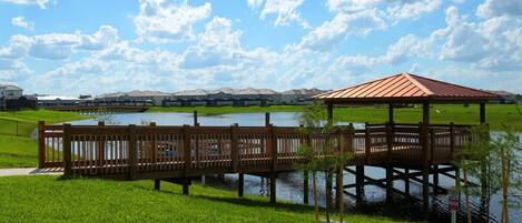 A wooden gazebo over a lake with a grassy foreground and a row of houses in the background.