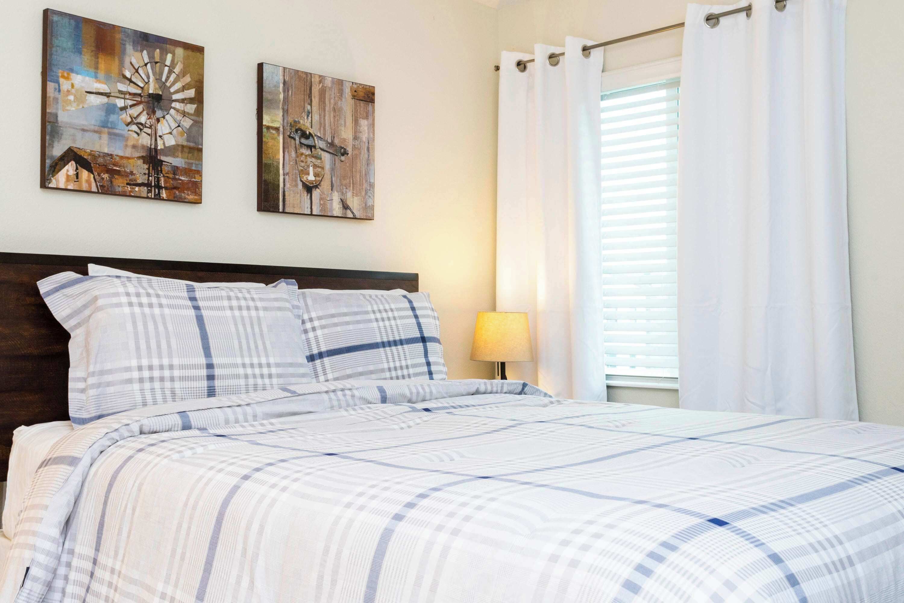 A neatly made bed with a striped bedspread, two wall-mounted paintings, a bedside lamp, and white curtains on a window.