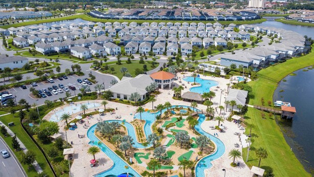 An aerial view of a residential area with a central swimming pool complex, surrounded by single-family homes, and a body of water to the right.