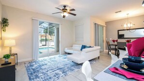 A modern living room with a white sofa, a dining table with chairs, a ceiling fan, and a view of the outdoors.