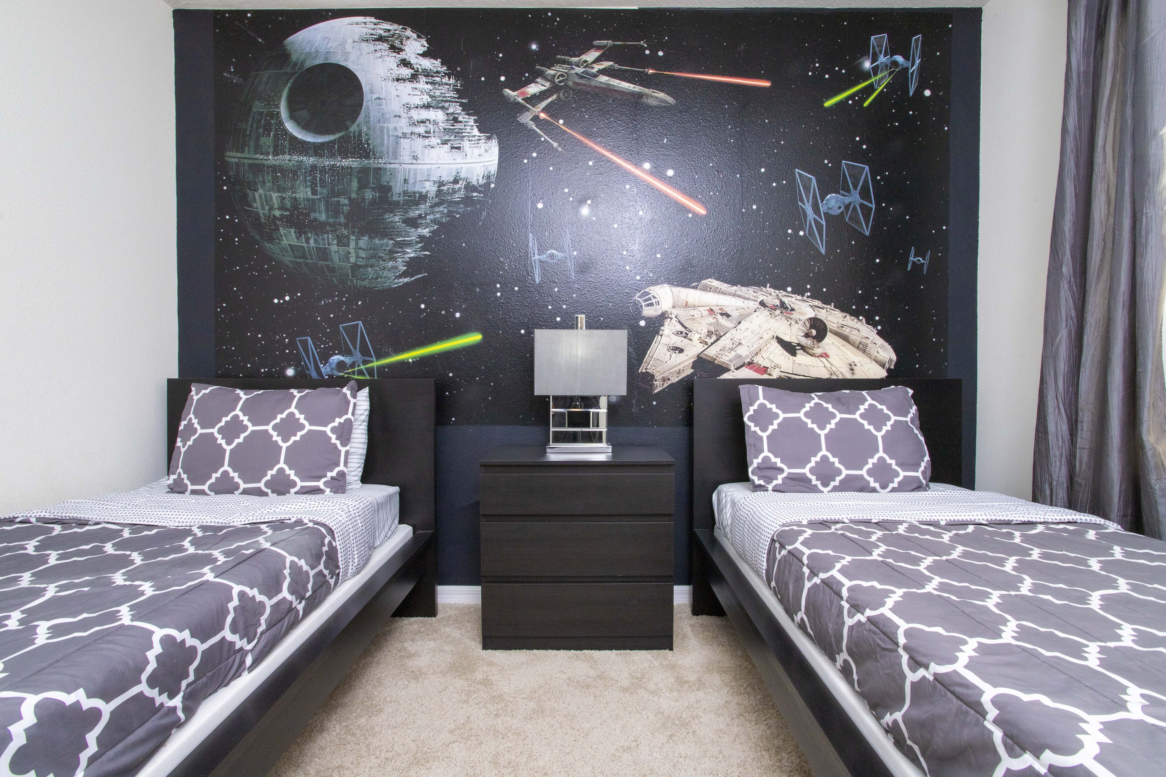 Une chambre avec deux lits, un mur décoré sur le thème de Star Wars, une table de chevet et une lampe.