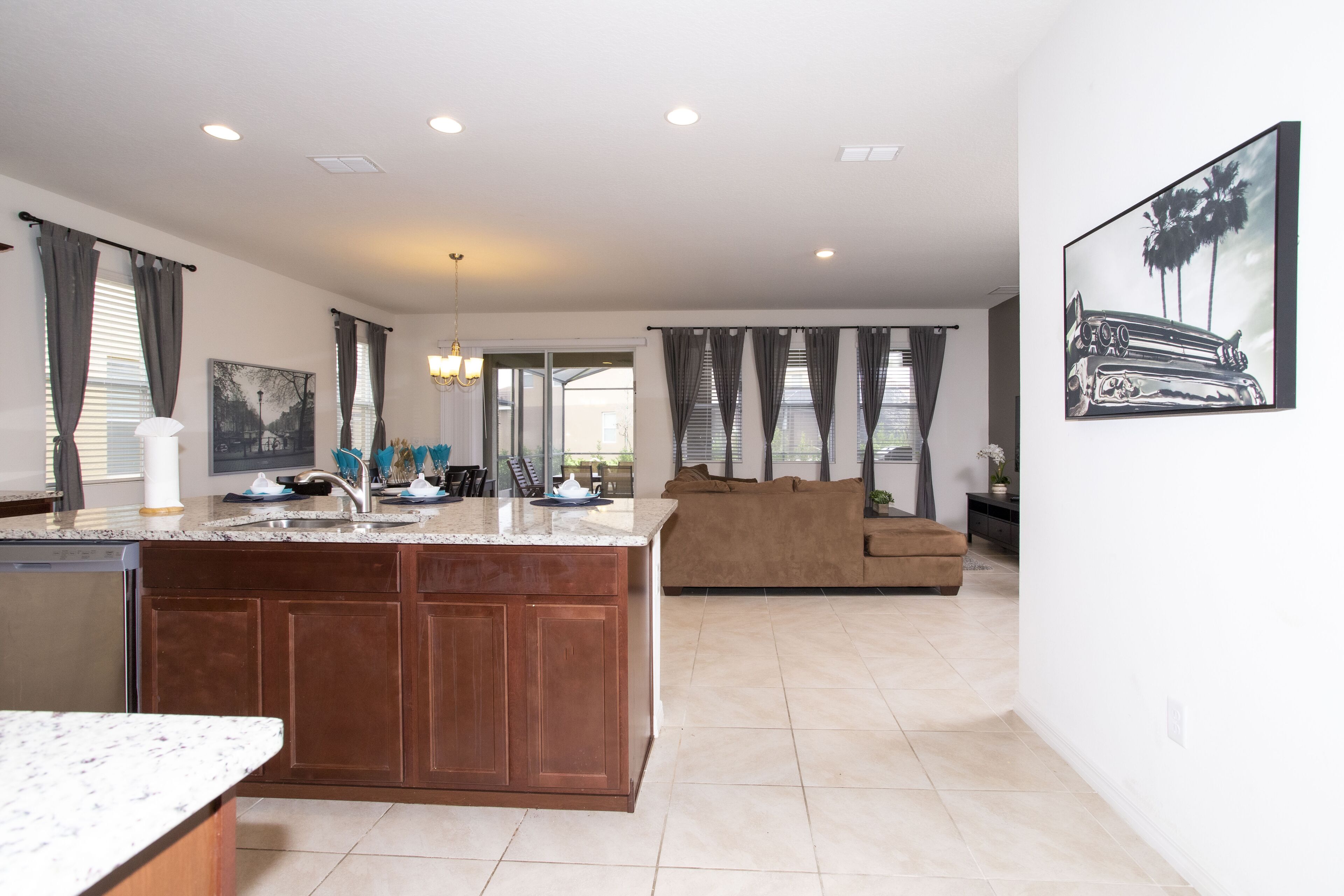 A modern kitchen with a central island, a dining area with a table and chairs, a living room with a sofa, and a large window with curtains.