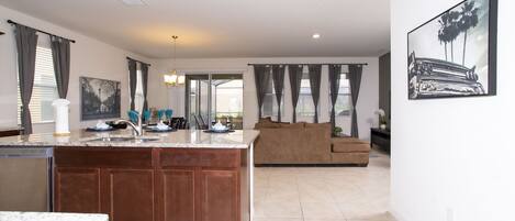 A modern kitchen with a central island, a dining area with a table and chairs, a living room with a sofa, and a large window with curtains.