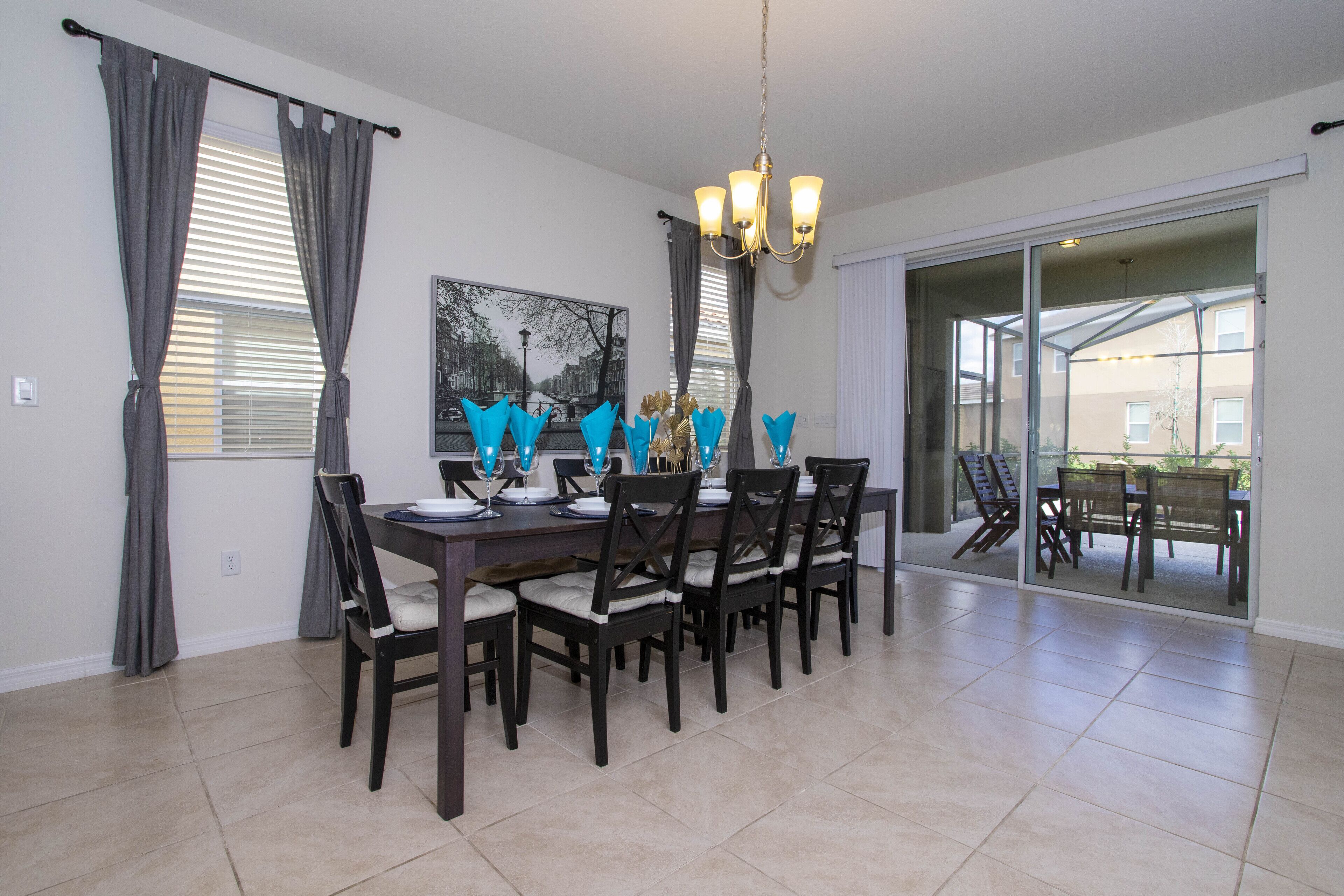A dining area with a table set for six, six chairs, a chandelier, and a large window with curtains.