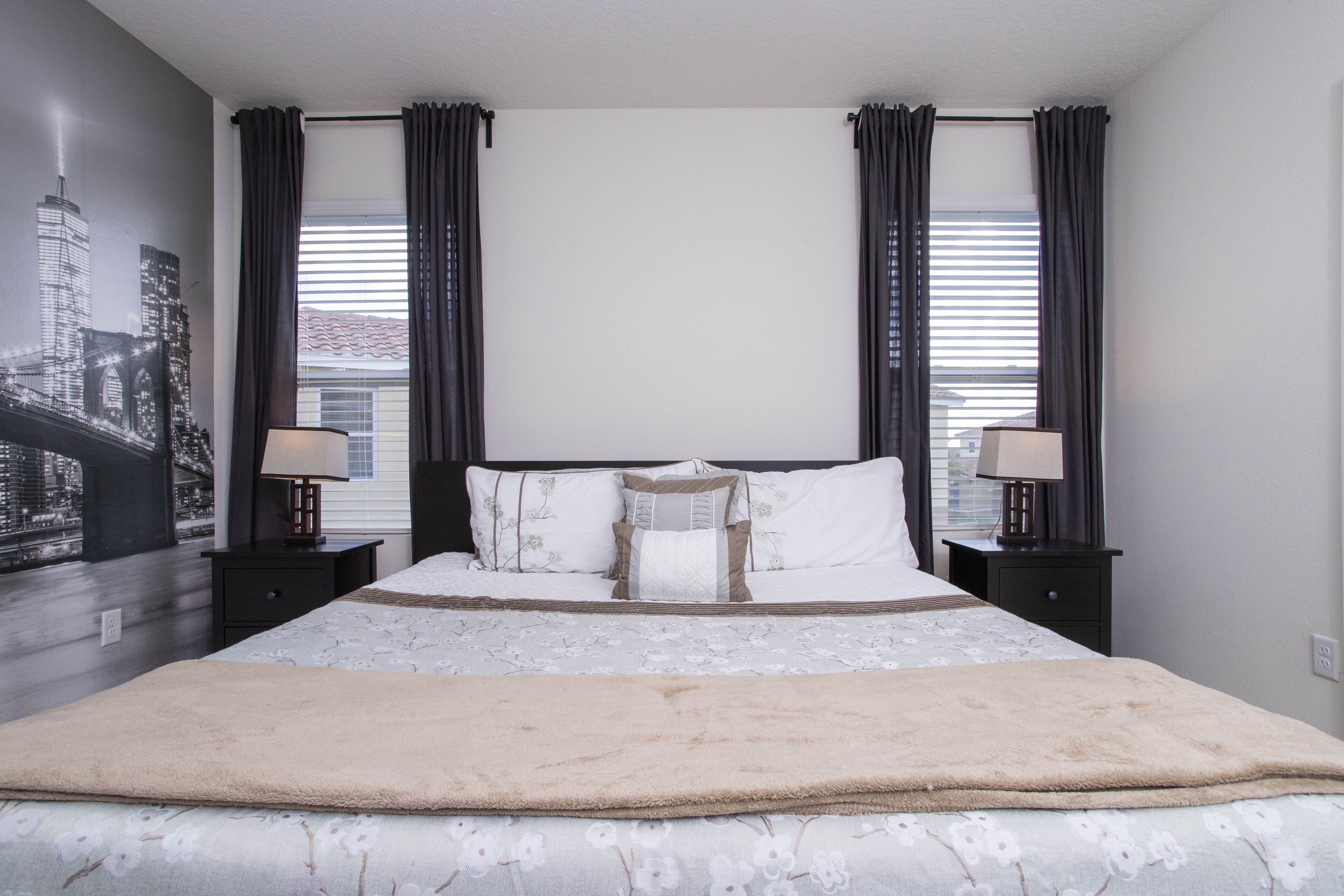 A bedroom with a large bed, two bedside tables with lamps, a cityscape mural on the wall, and a view of the city through the window.