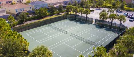 A tennis court surrounded by residential buildings, trees, and a parking area.
