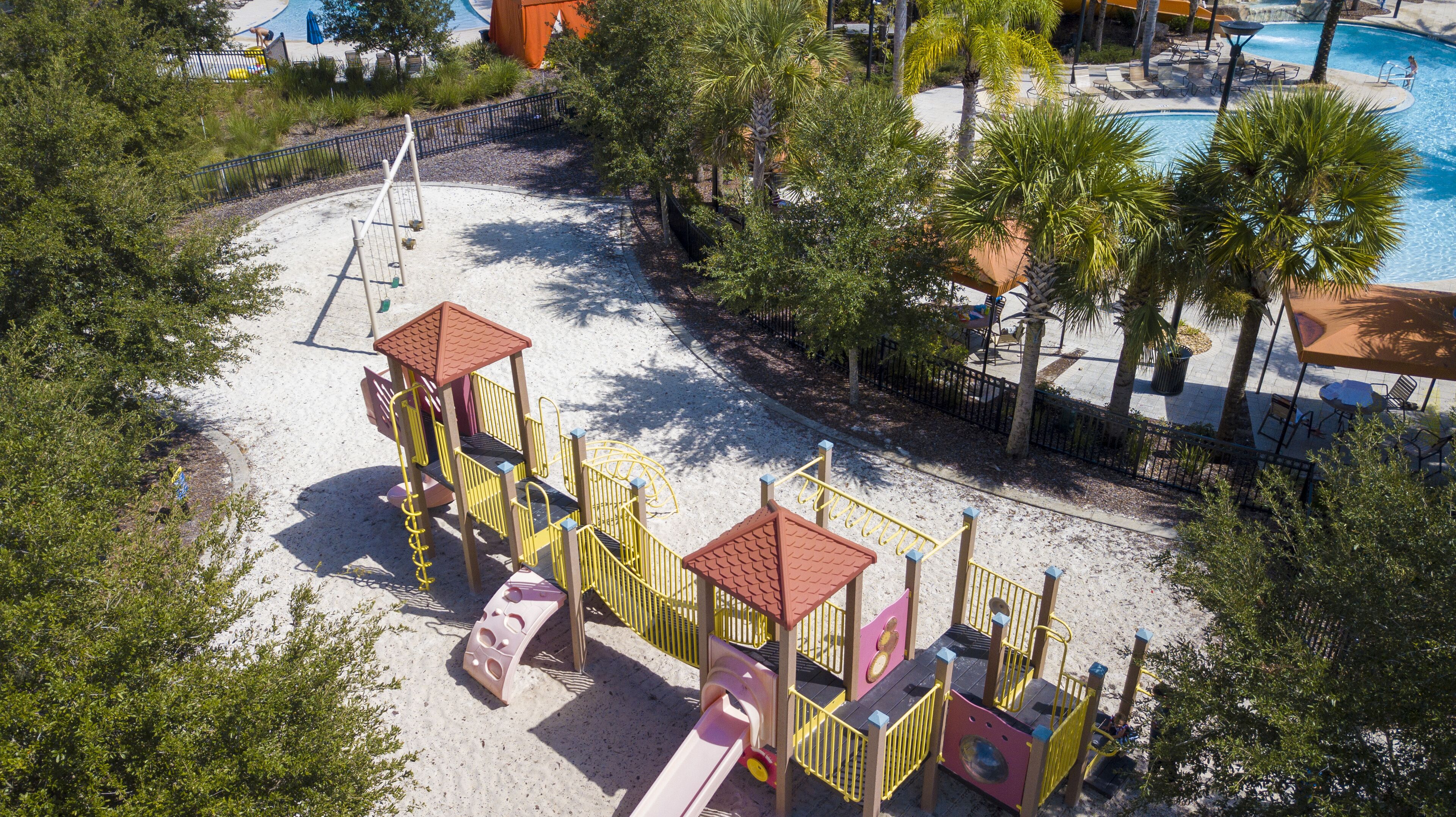 A playground with slides and climbing structures, surrounded by palm trees and a pool area.
