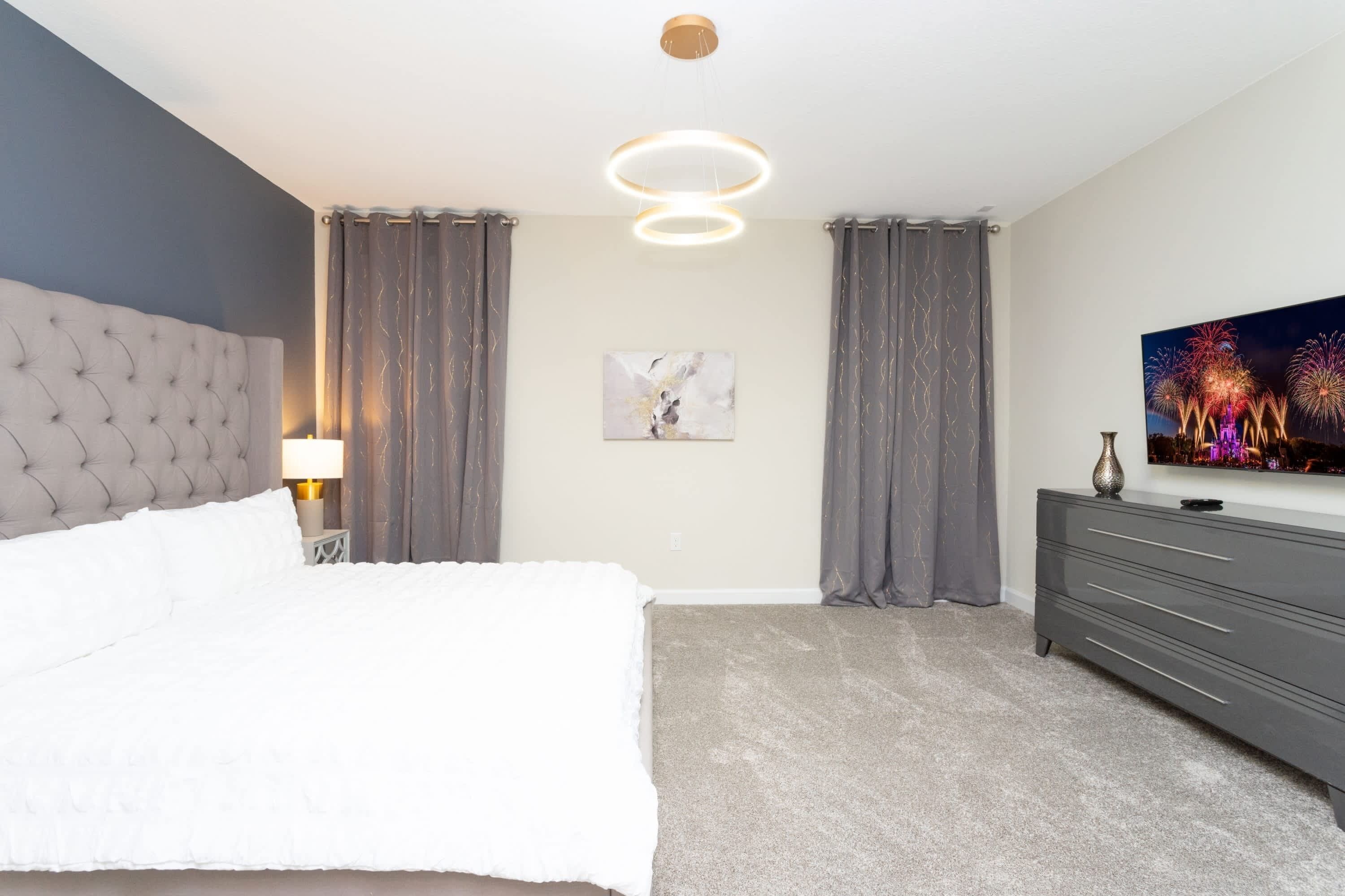 A modern bedroom with a tufted headboard, a bed with white bedding, a bedside lamp, grey curtains, a grey dresser, a framed abstract painting, and a wall-mounted TV displaying fireworks.