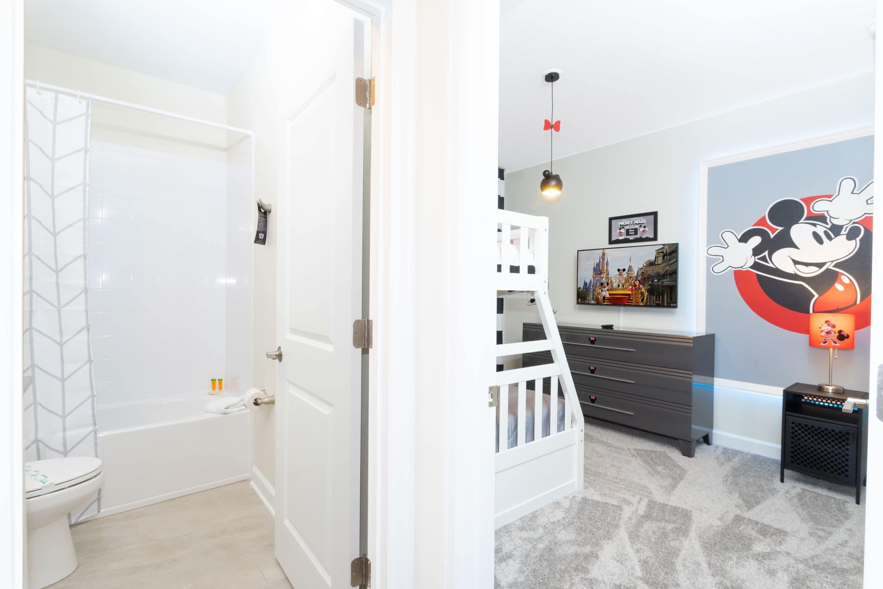 A child's room with a white door, a white crib, a dresser, a Mickey Mouse wall decal, and a lamp.
