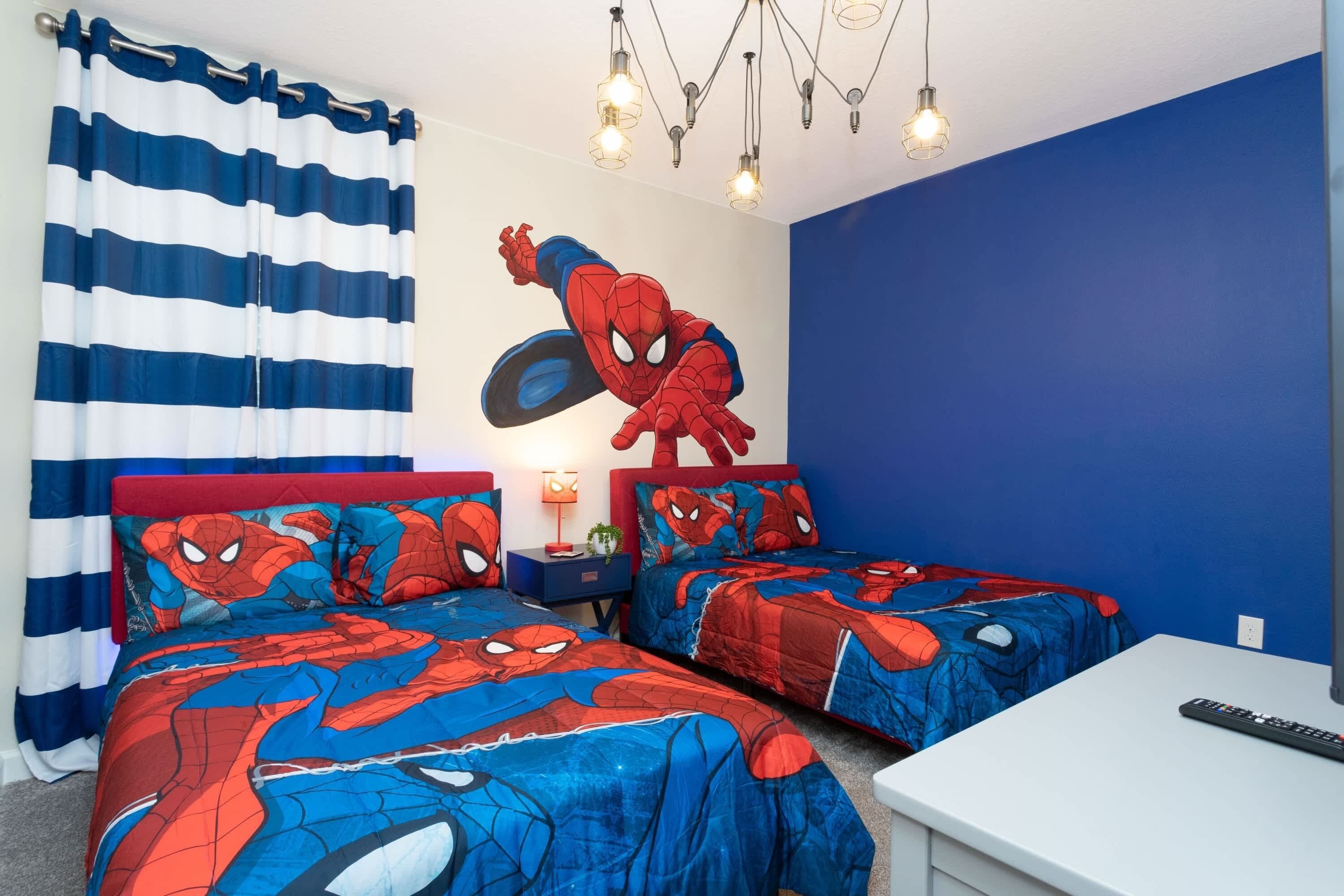 A bedroom with two beds, each with a Spider-Man themed bedspread, a nightstand with a lamp, a ceiling light fixture, and a TV on a stand.