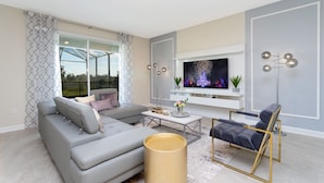 Een moderne woonkamer met een grijze modulaire bank, een klein rond tafeltje, een vloerlamp en een aan de muur bevestigde televisie.