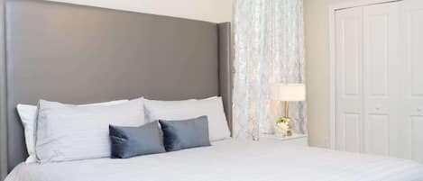 A neatly made bed with white and gray bedding, a bedside table with a lamp, and a patterned curtain.