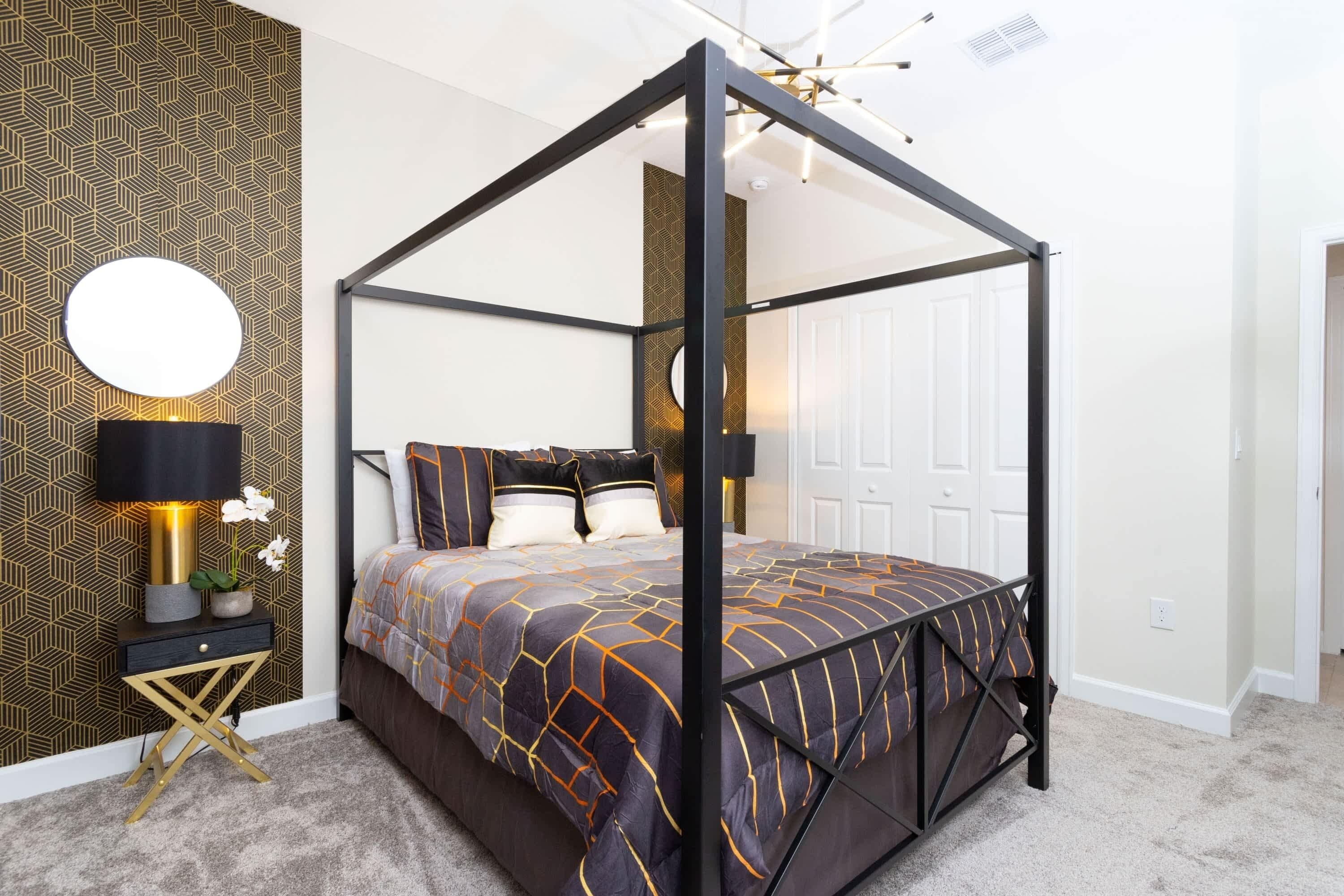 Ein Bett mit Baldachin, dessen Bettdecke ein geometrisches Muster aufweist, ein Nachttisch mit Lampe sowie eine Wand mit geometrischer Tapete.