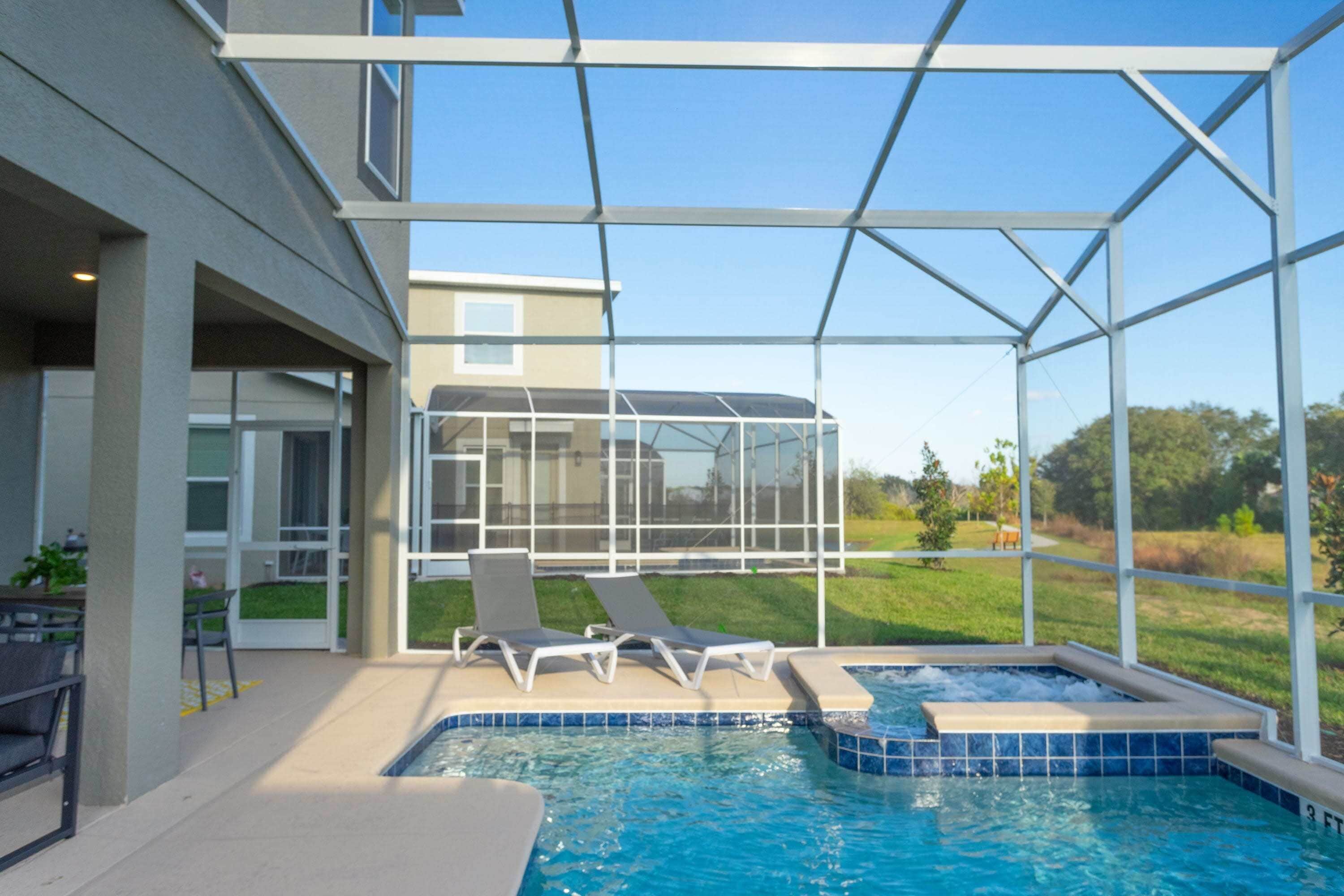 Une piscine entourée de parois en verre, deux chaises longues et un espace patio avec une table et des chaises.