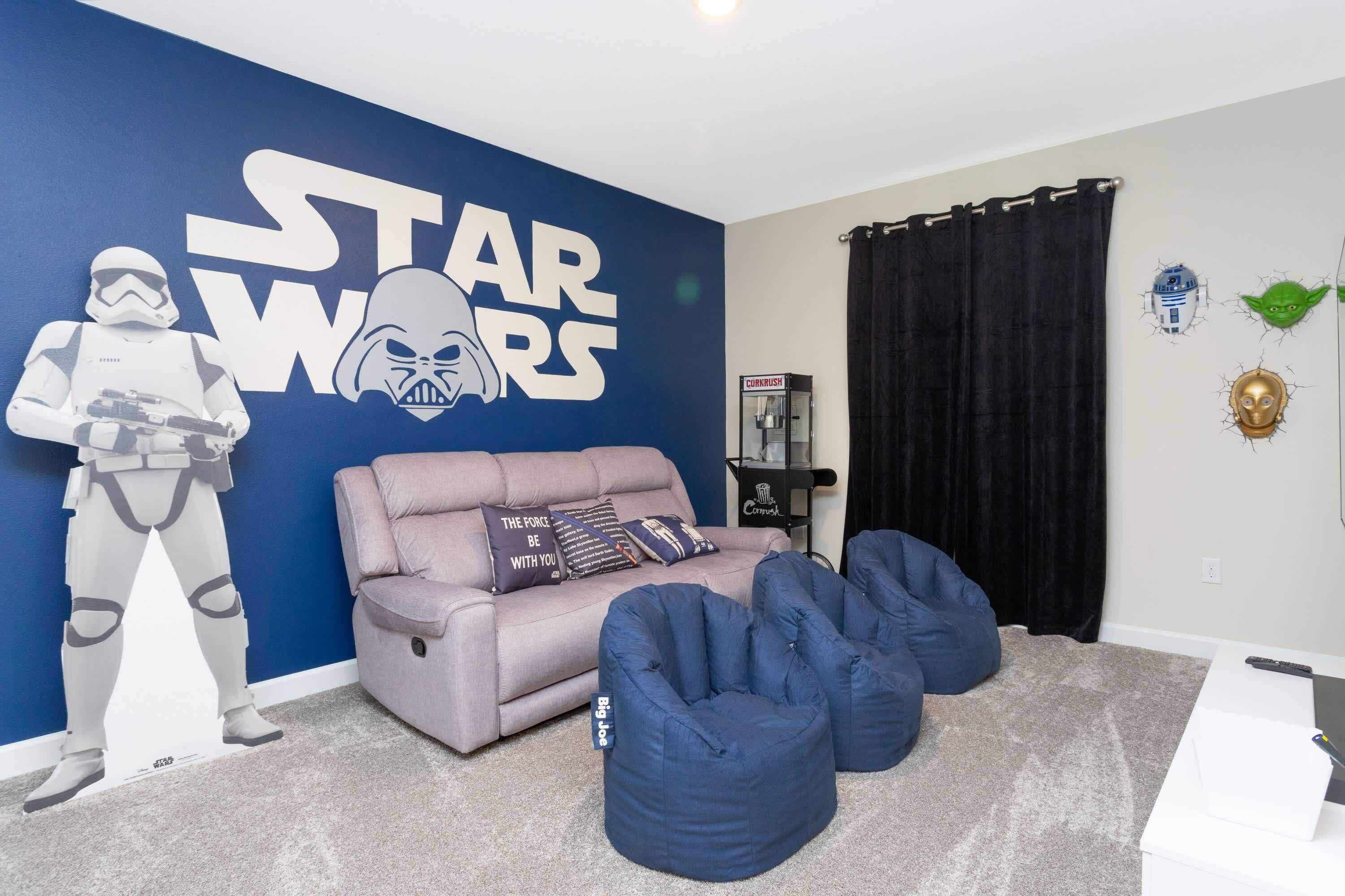 Una habitación con una pared temática de Star Wars, un sofá gris con almohadas y pufs azules sobre un suelo alfombrado.