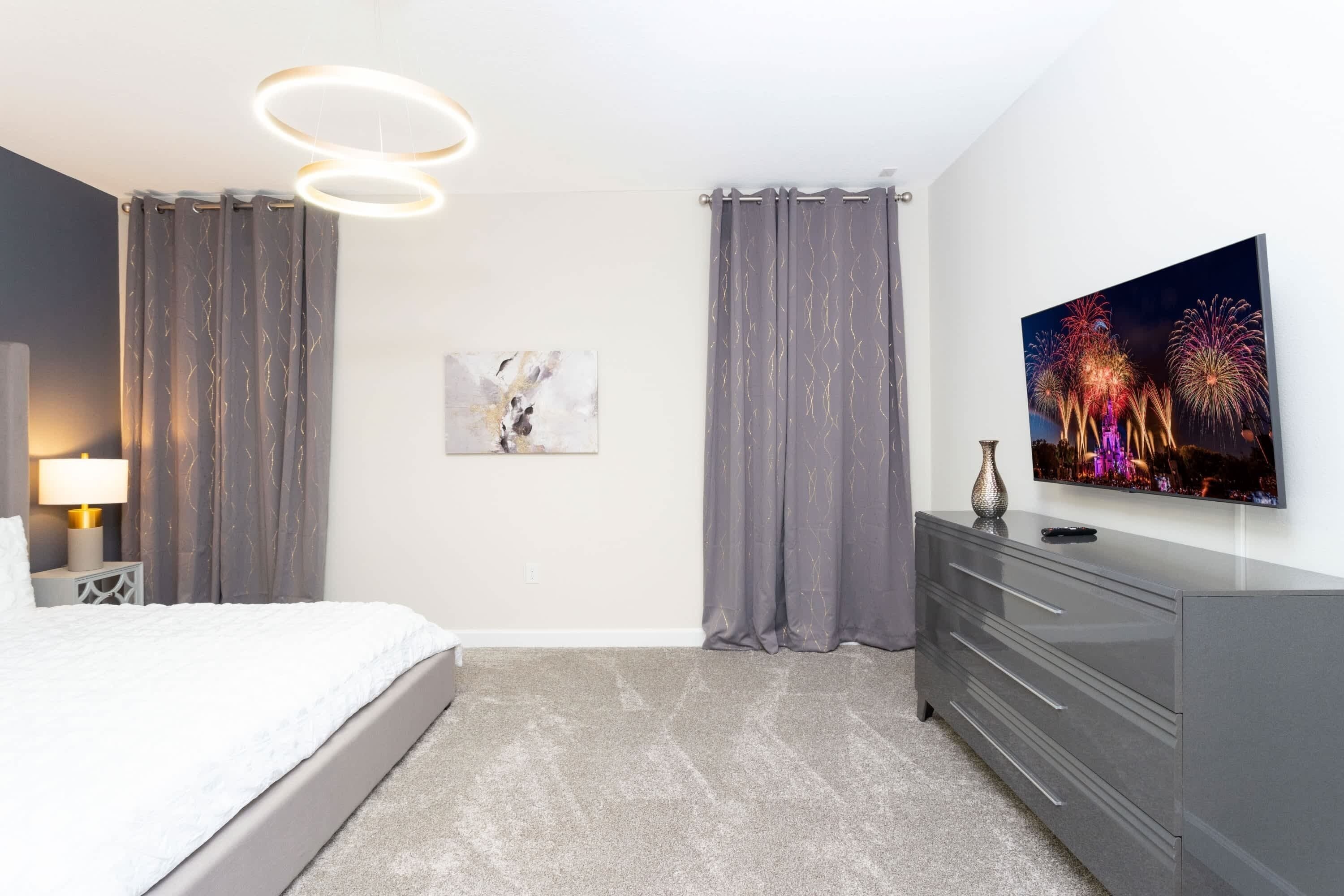Un dormitorio moderno con cama, cortinas grises, televisor de pantalla plana, cómoda y lámpara fijada a la pared.