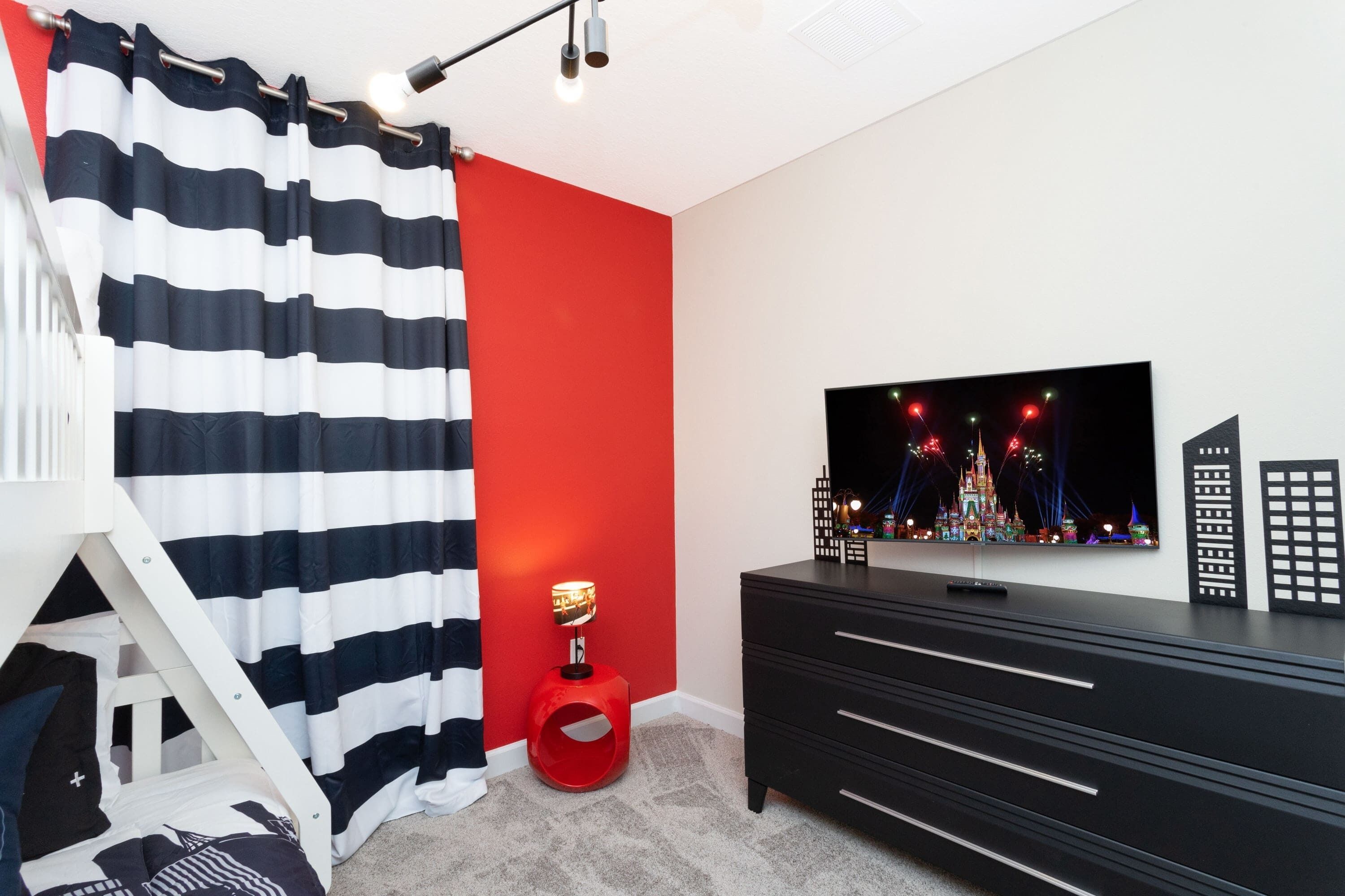 A room with a TV on the wall, a red accent wall, a black dresser, and a striped curtain.