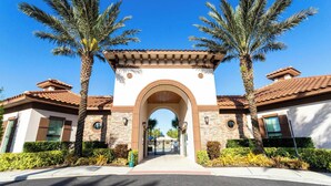 A gated entrance with a central archway, flanked by palm trees and a clear blue sky. - Stunning 5Bd w/ Pool Close to Disney @ Solterra 7829 (Davenport)