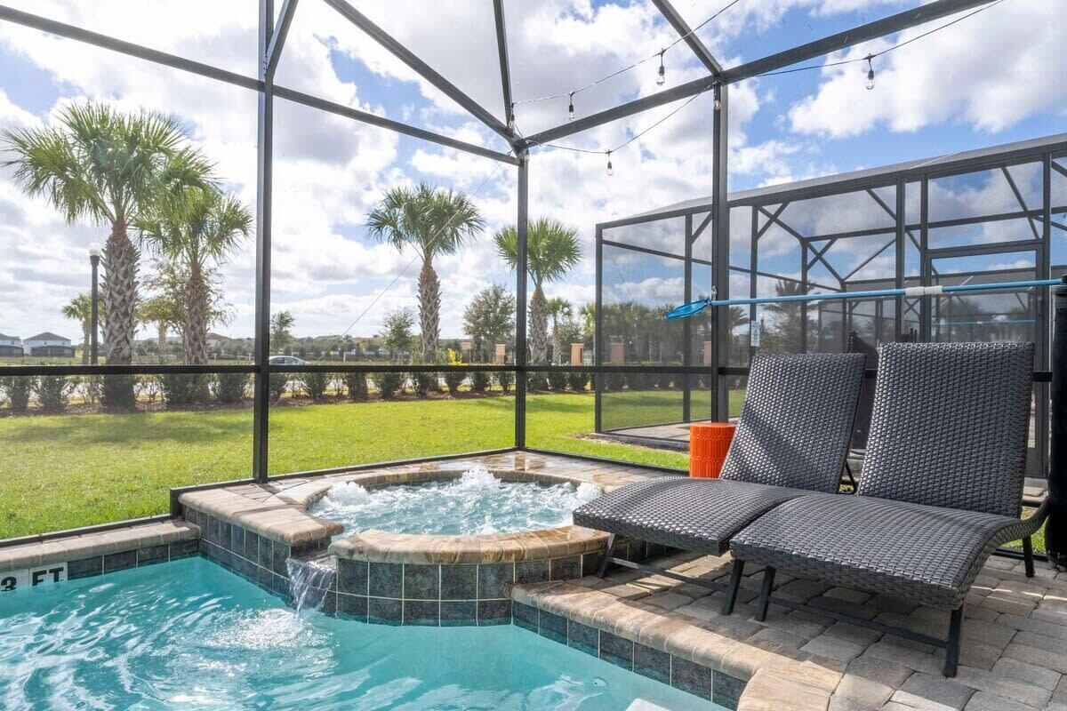 A covered outdoor swimming pool with a hot tub, lounge chairs, and a view of palm trees and a grassy area.