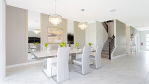 Uma sala de jantar espaçosa com piso de mármore, cadeiras brancas e uma grande mesa. Dois candelabros pendem do teto.