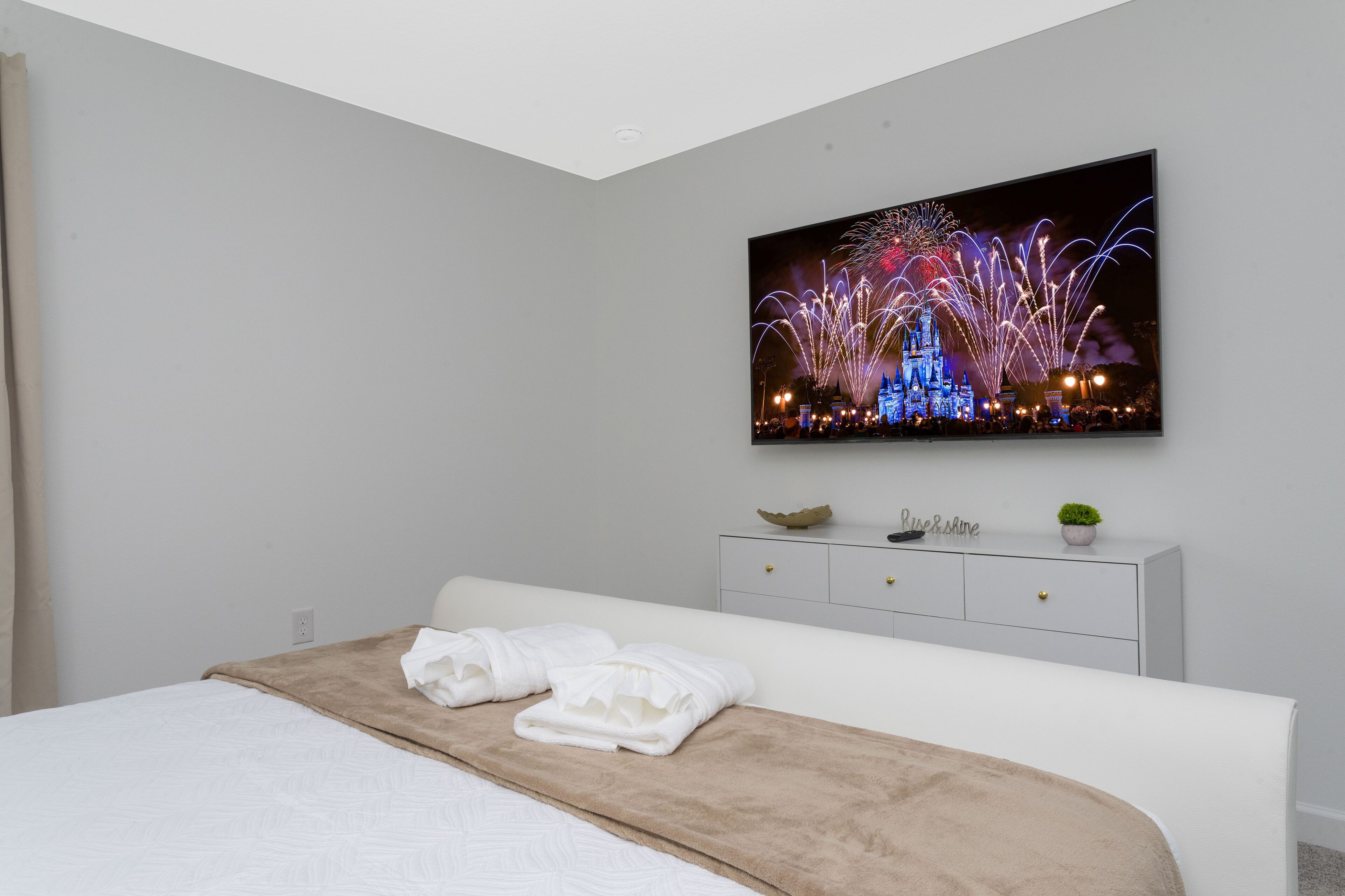 Une chambre avec un lit, une commode blanche, un téléviseur à écran plat fixé au mur et une petite plante posée sur la commode.