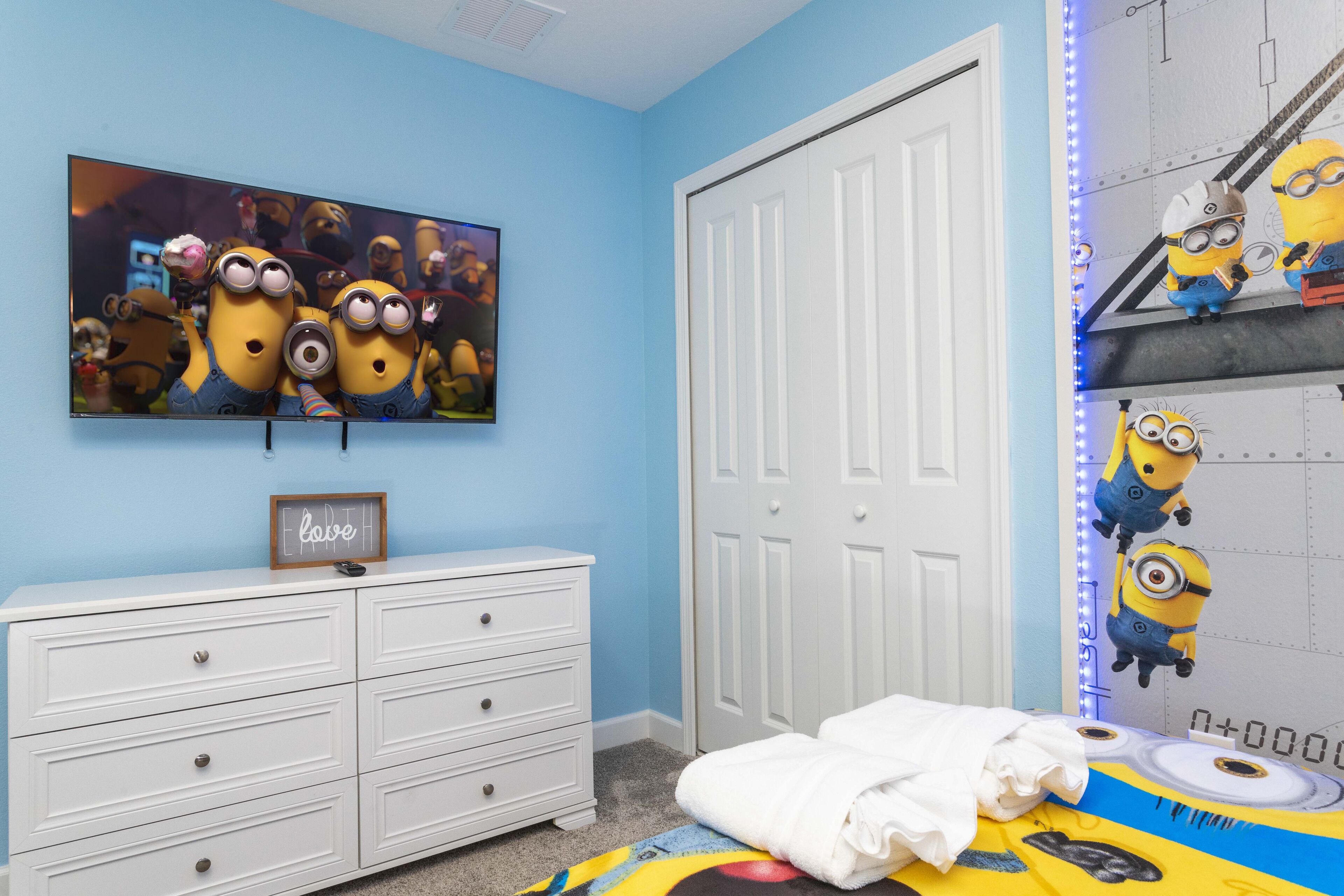 Un dormitorio con pared azul, cómoda blanca, cama con manta temática de Minions, un cuadro enmarcado sobre la cómoda y estante con Minions en la pared.