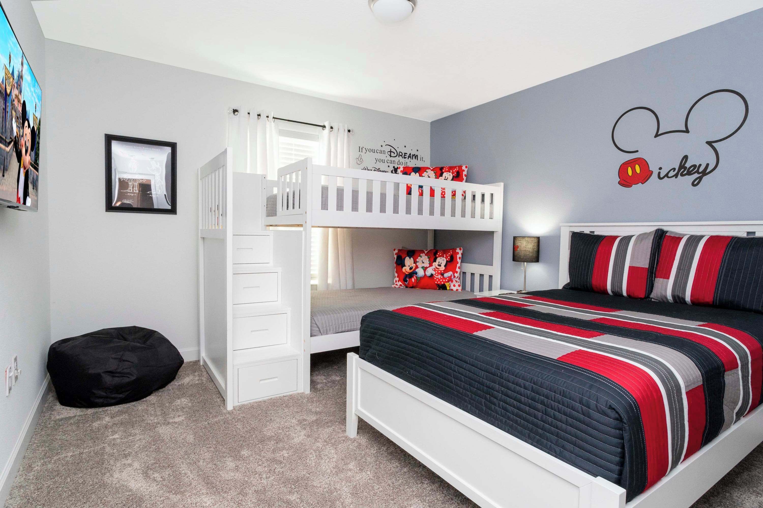 Une chambre d’enfant avec des lits superposés, un lit recouvert d’une couverture à rayures, un autocollant mural représentant Mickey Mouse et un tableau encadré accroché au mur.