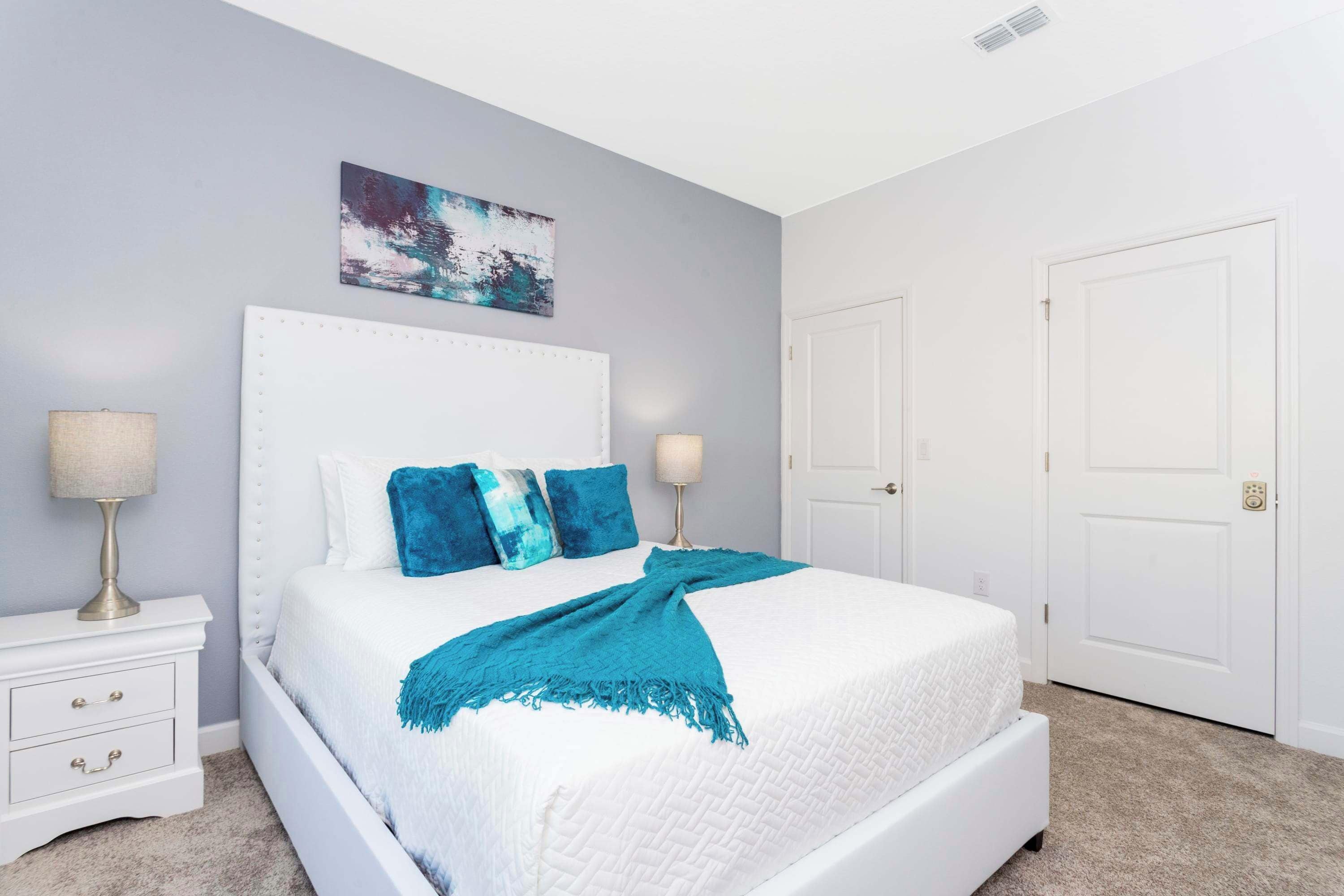 A bedroom with a white bed, blue pillows, a white headboard, and a teal throw blanket. There are two bedside tables with lamps and a door to the right.