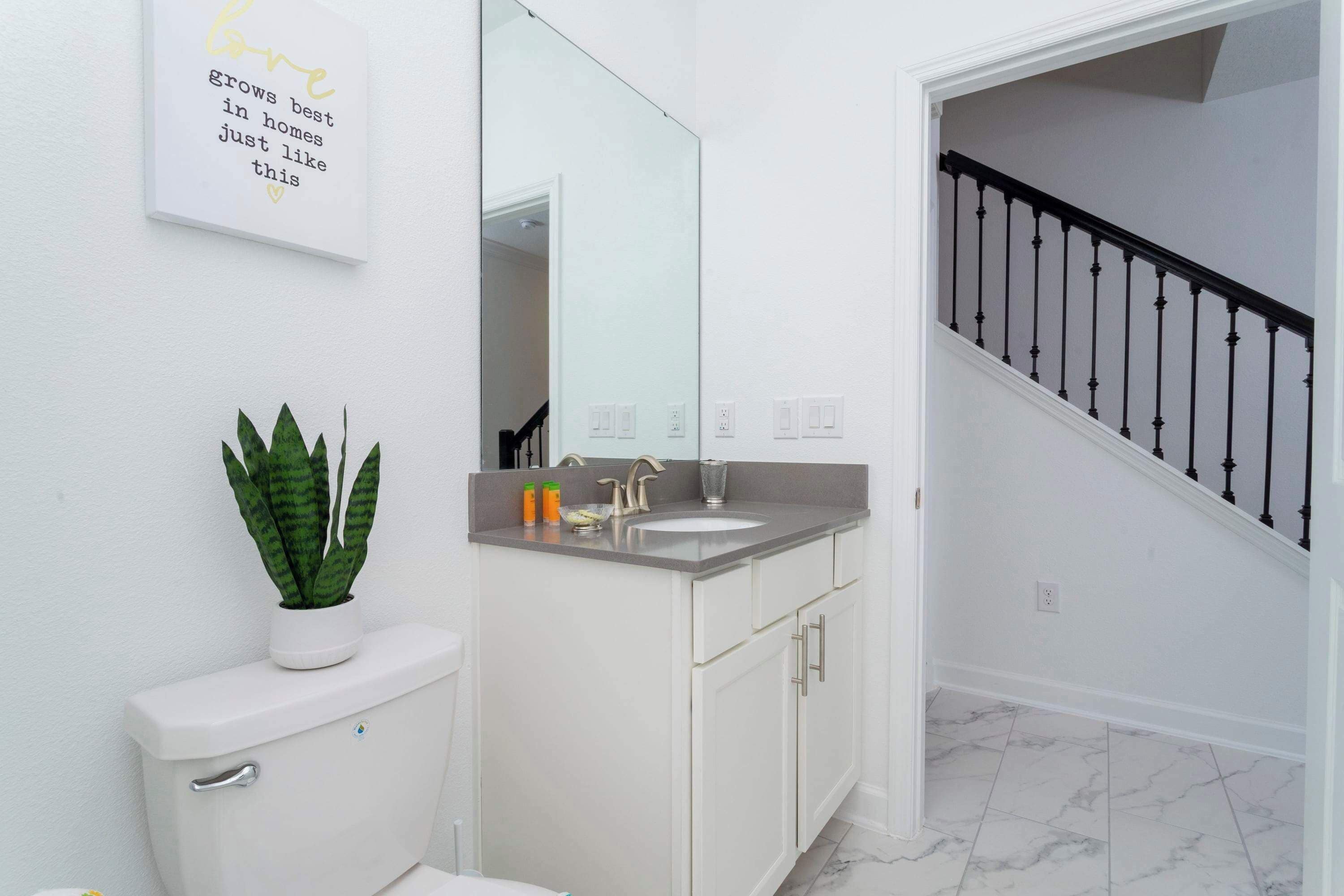 A bathroom with a toilet, sink, and mirror. There is a staircase with a black railing leading to another room.
