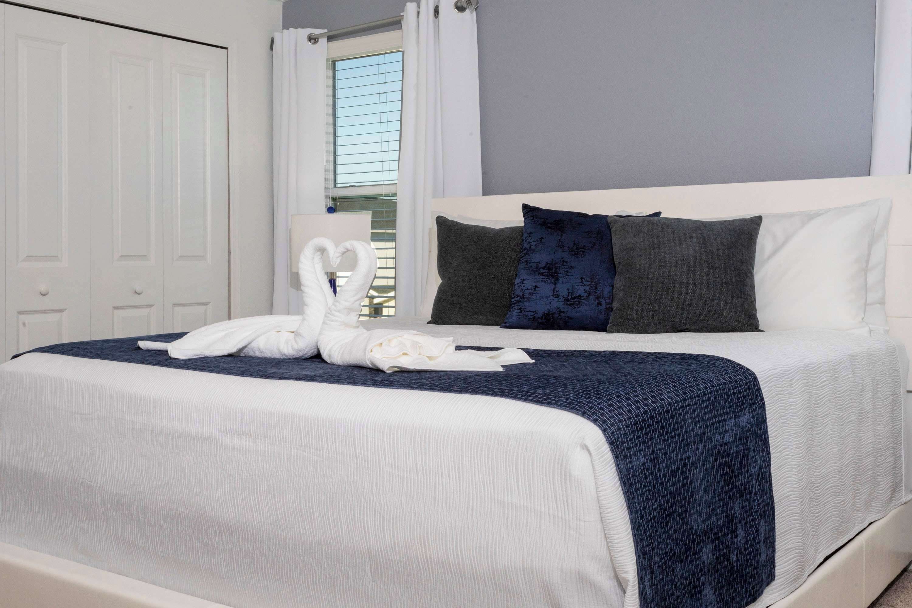 A neatly made bed with a white and blue bedspread, two pillows, and a towel arranged in a heart shape.