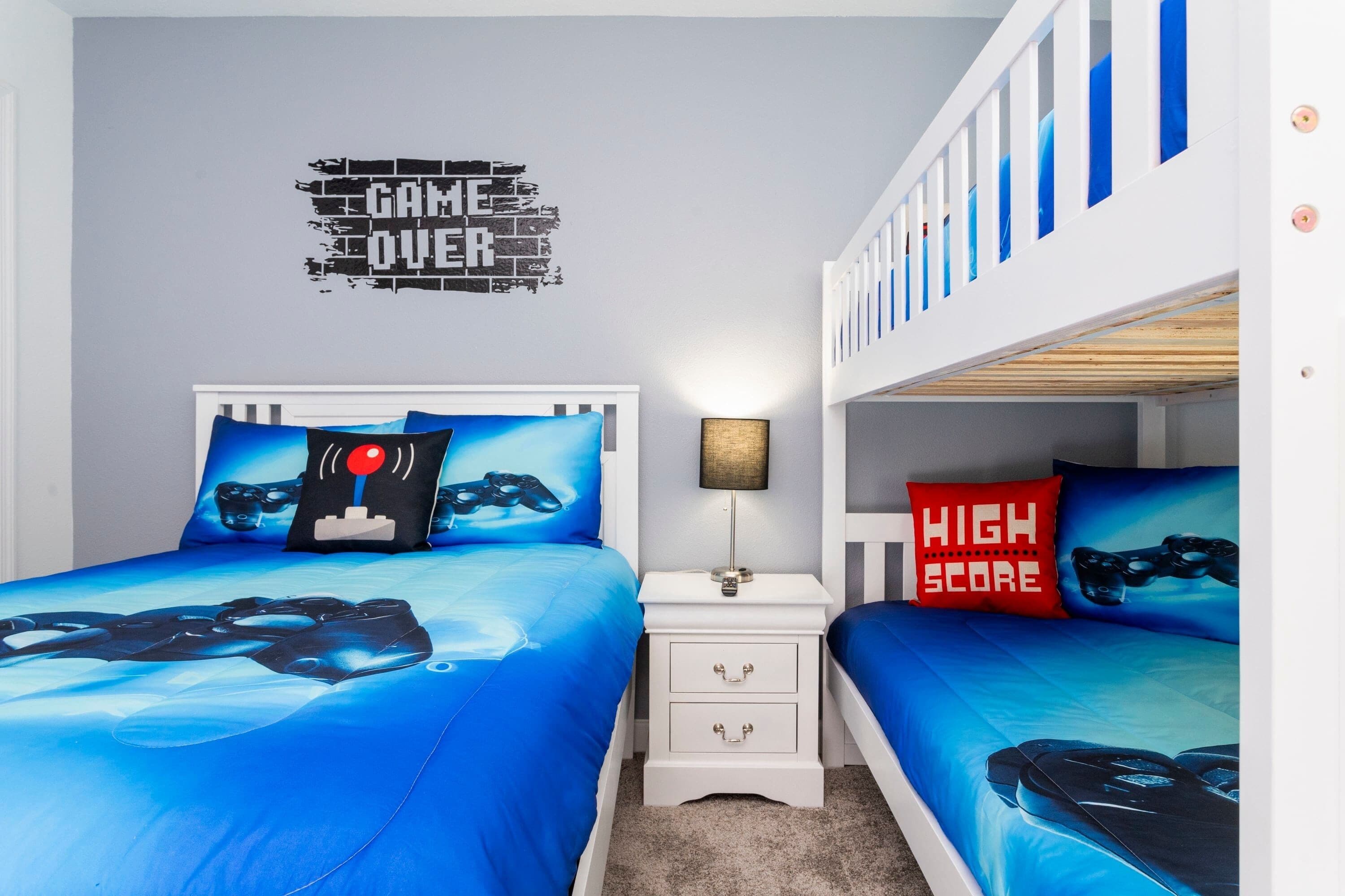 Ein Schlafzimmer mit zwei Betten, beide mit blauer Bettdecke mit einem Design von Spielecontrollern. An der Wand befindet sich ein Wandbild mit der Aufschrift 'GAME OVER' und ein kleiner Tisch mit einer Lampe zwischen den Betten.
