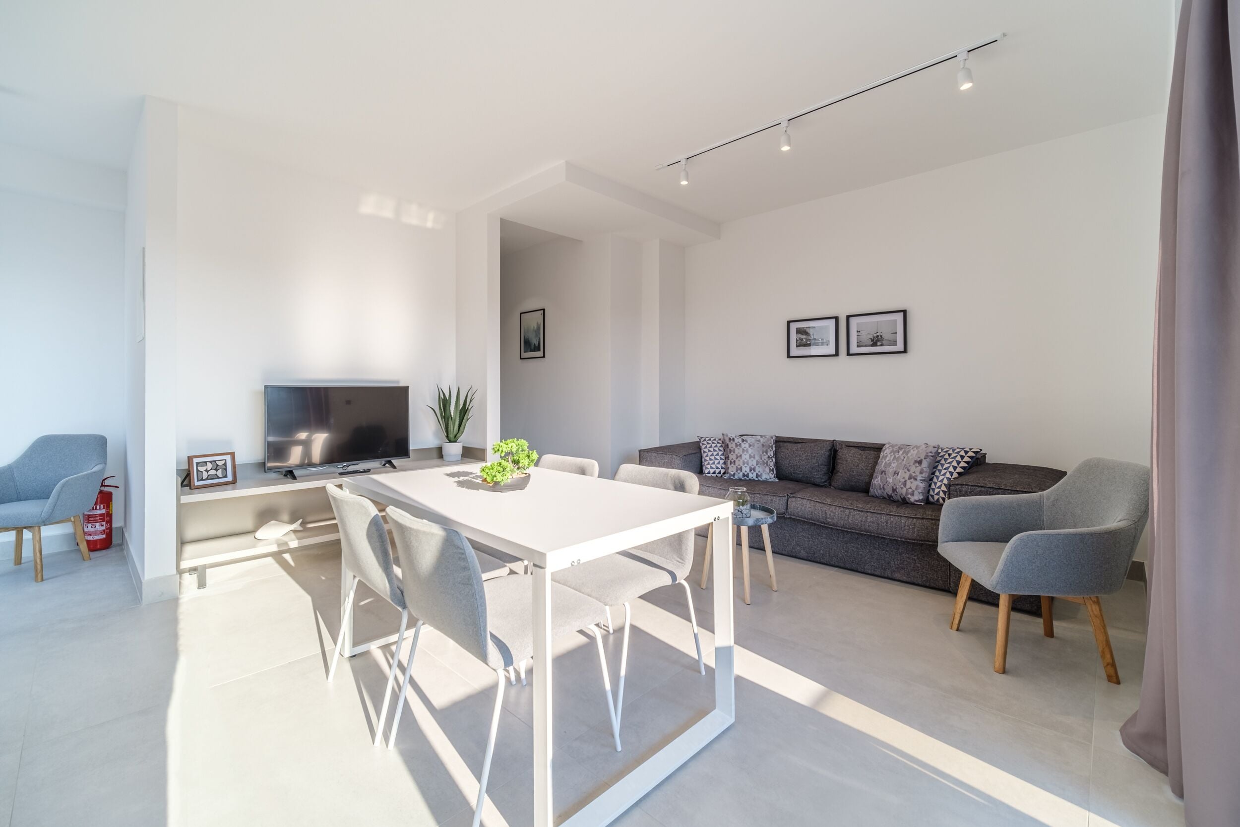 A modern living room with a white dining table, grey sofa, and a flat-screen TV.