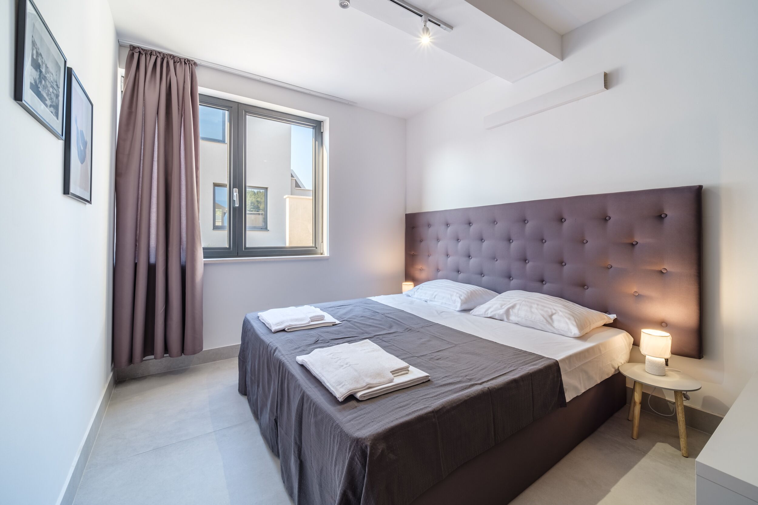 A modern bedroom with a large bed, a headboard, a bedside table with a lamp, and a window with curtains.