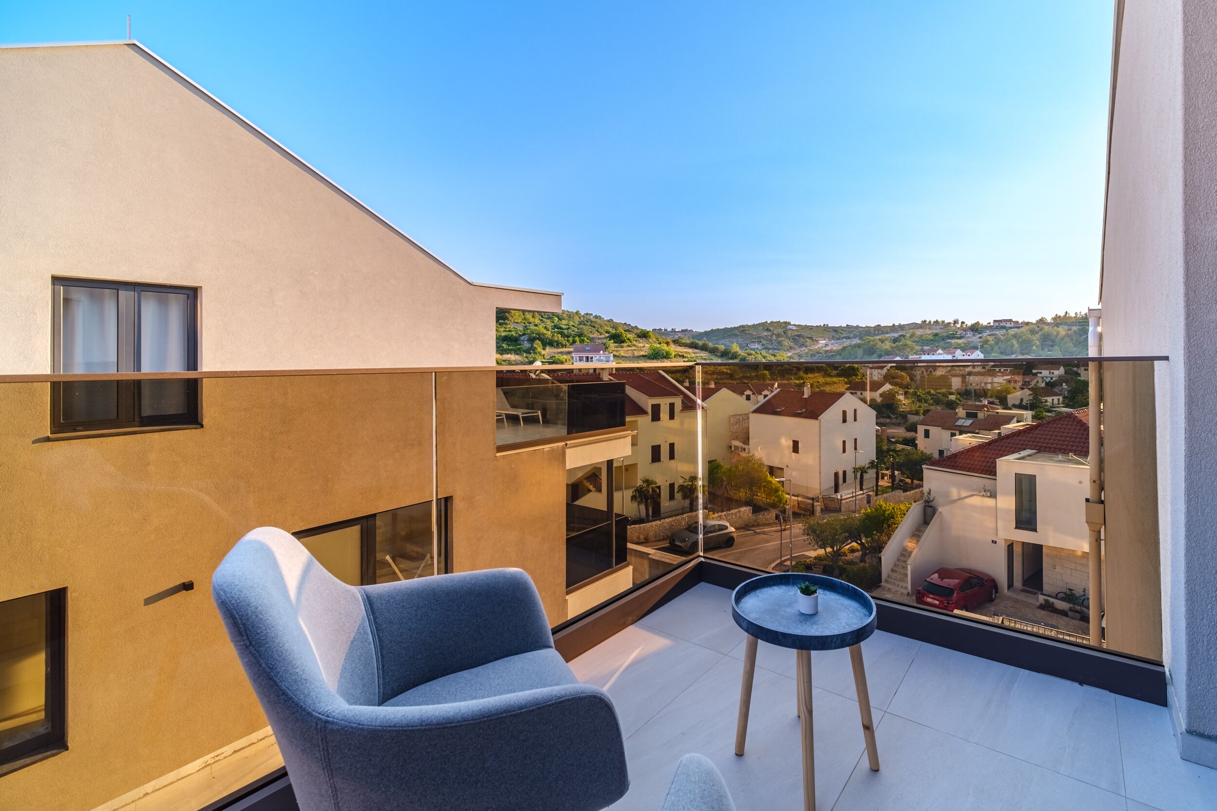 A balcony with a blue chair and a small table, overlooking a residential area with houses and hills.
