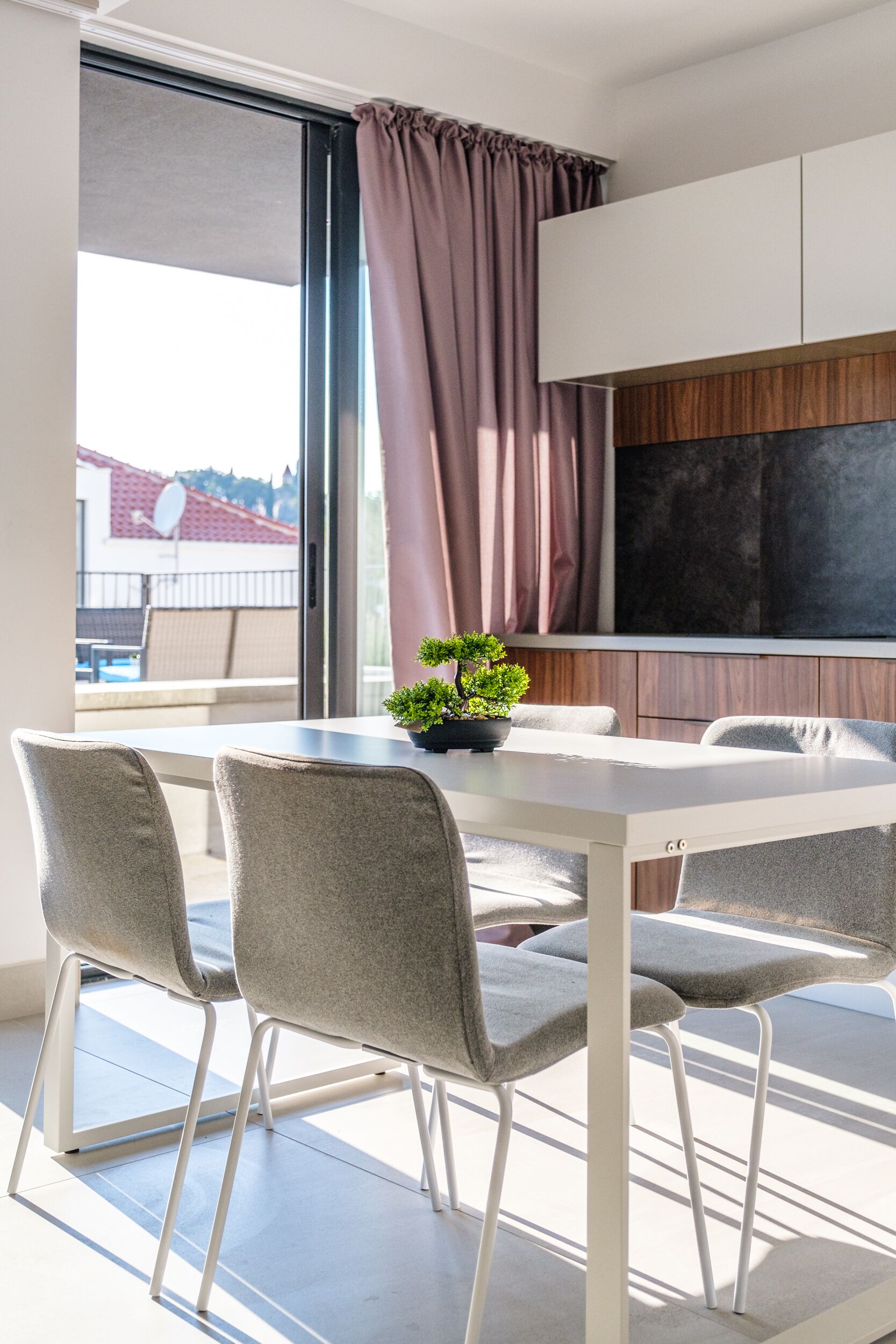Et moderne spiseområde med et hvidt bord, fire grå stole, en potteplante og udsigt til en altan med rødt tag.