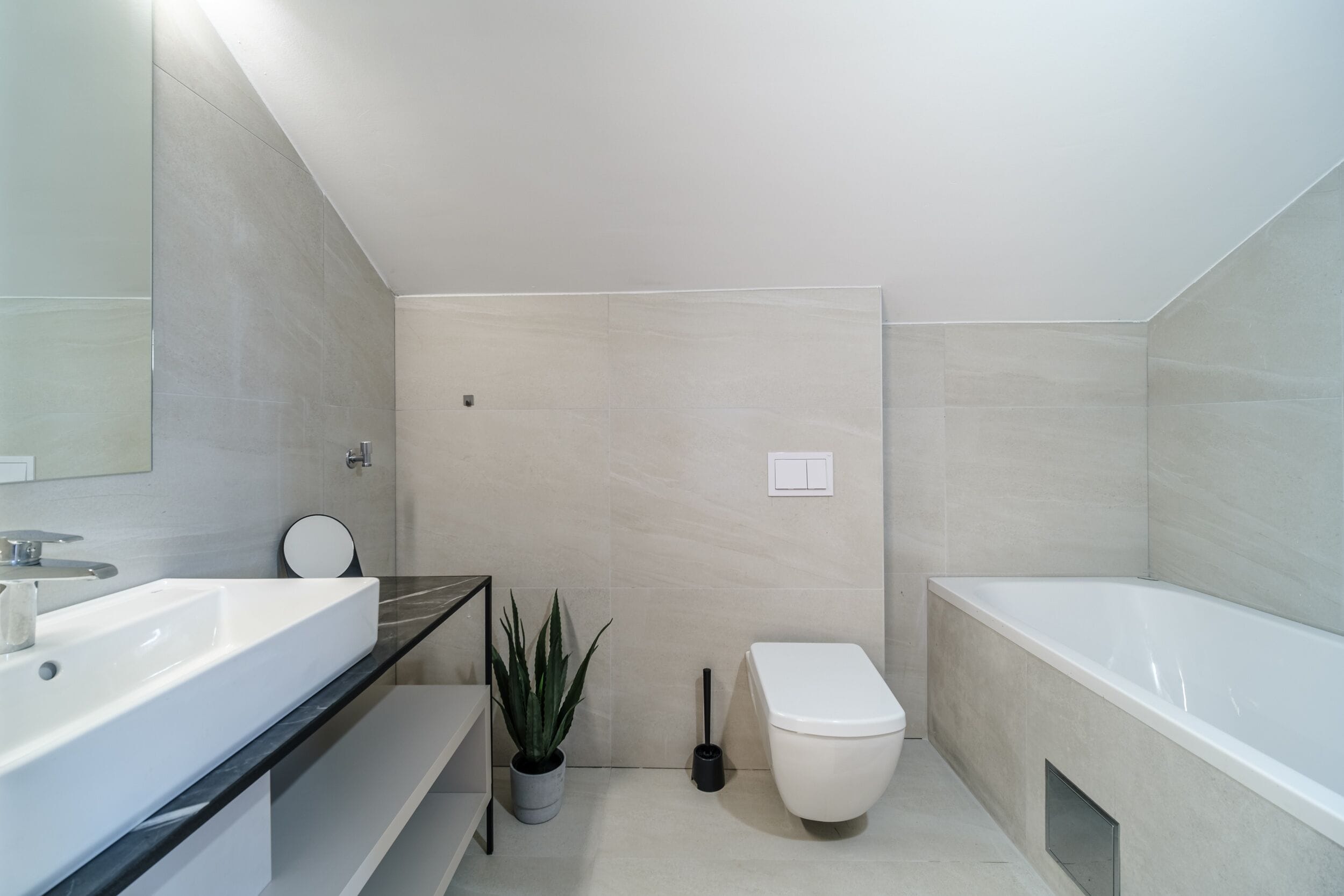 A modern bathroom with a large white bathtub, a white sink, and a toilet. There is a potted plant and a black cleaning brush on the floor.