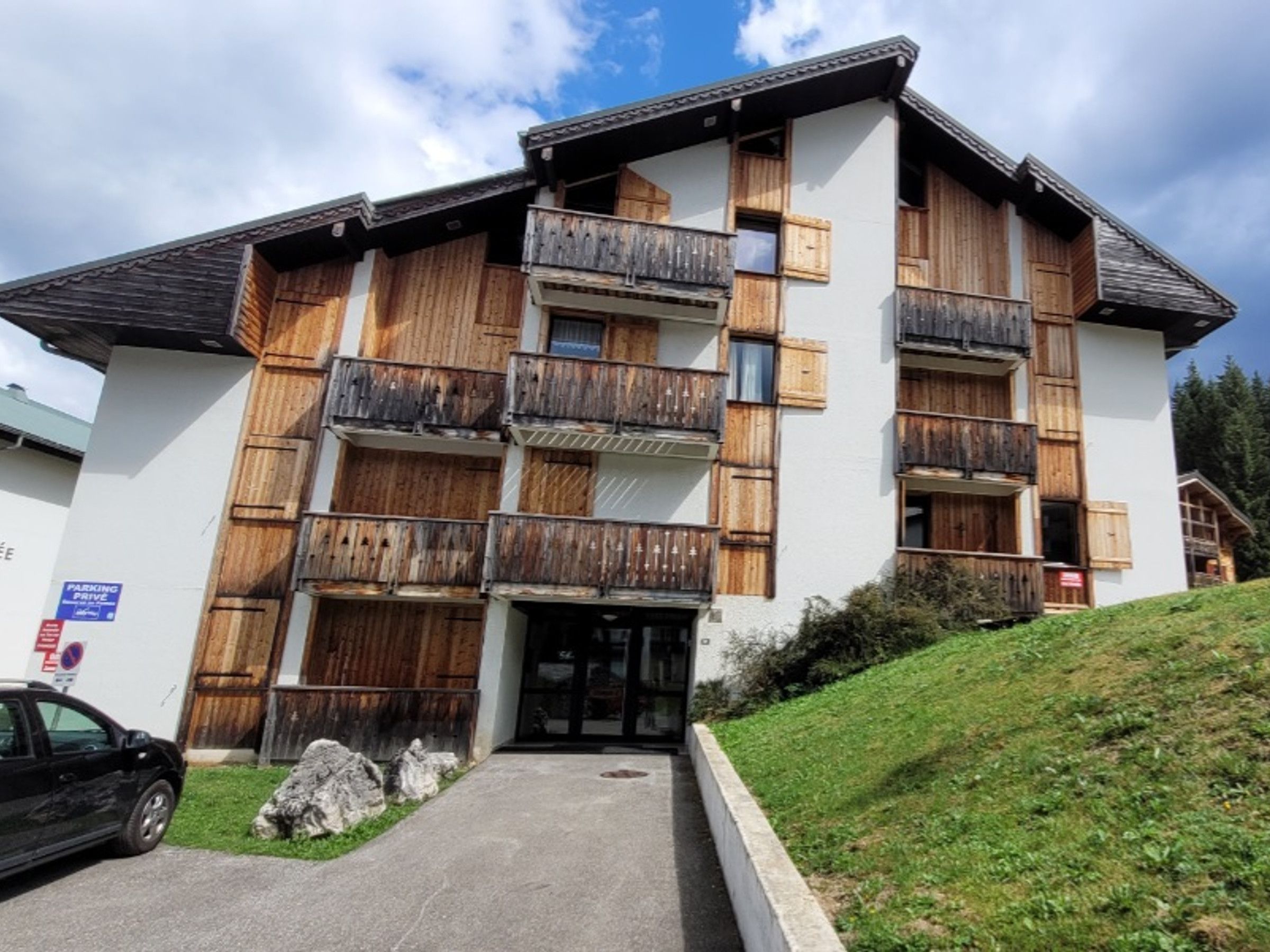 A multi-story building with wooden balconies, a paved entrance, and a car parked in front.