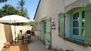 A patio area with a wooden table and chairs, a white umbrella, and a welcome sign on a building with green shutters.
