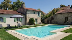 A rectangular swimming pool with a surrounding deck, adjacent to a building with a red-tiled roof and a stone facade.